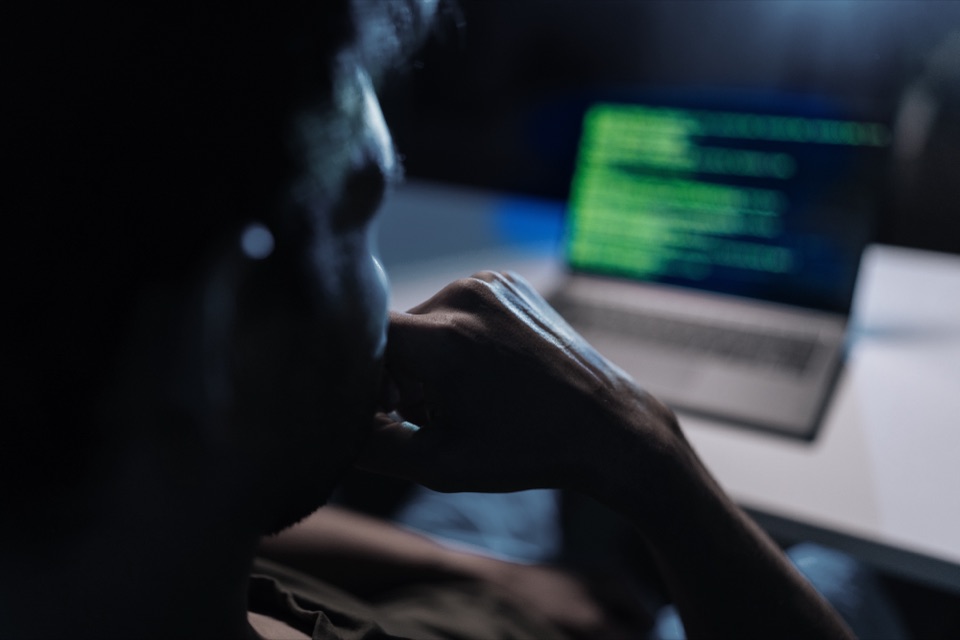Person sitzt im Dunkeln vor einem Laptop, auf dessen Bildschirm gr&uuml;ner Programmcode leuchtet
